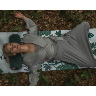 Kobieta leży na macie pośród liści w lesie, pod głową ma mały wałek gryczany WellB w butelkowej zieleni.